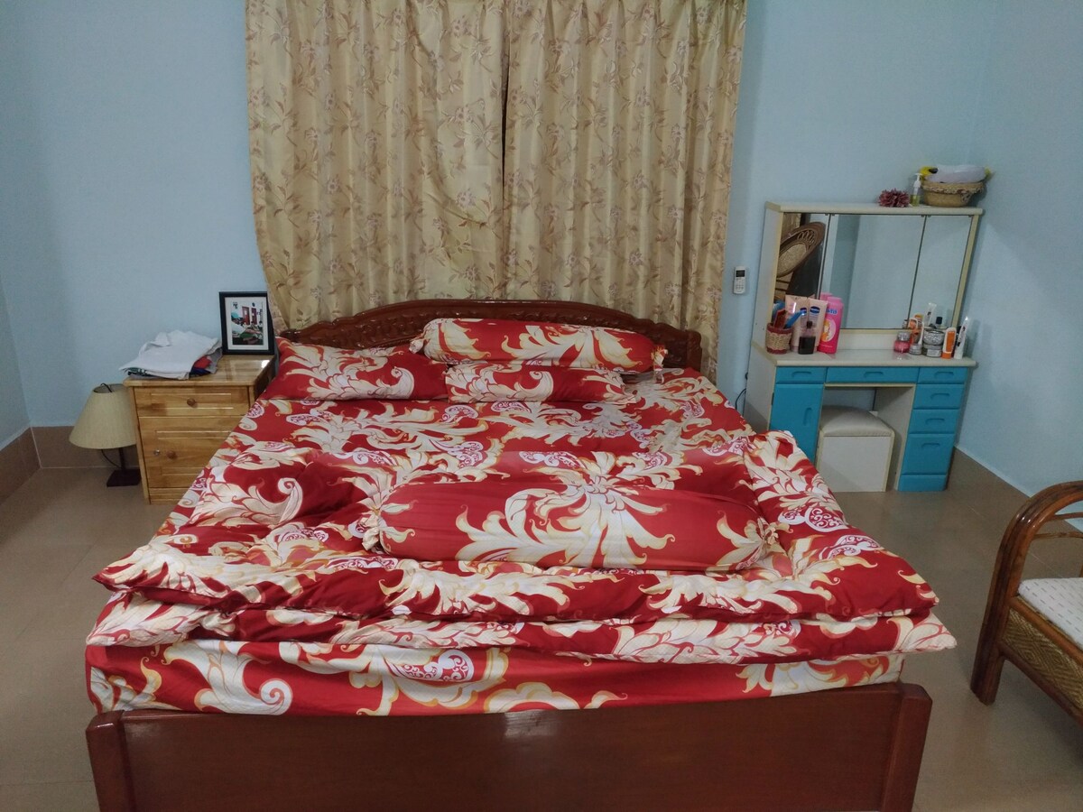 The bedroom features a wooden bed adorned with a vibrant red and gold patterned bedding set. A pair of bedside lamps are positioned on either side of the bed. A small dresser with a mirror and a blue vanity unit are visible against the light blue walls.