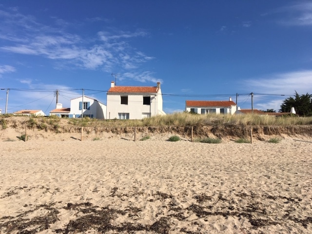 The image portrays a sandy beach with a gentle slope leading up to a grassy area. In the background, two white buildings with red roofs are visible, set against a clear blue sky. The calm atmosphere is highlighted by the simplicity of the beach environment.