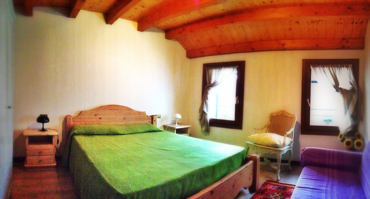 A spacious bedroom features wooden beams on the ceiling and light-colored walls. A large bed with a green coverlet is centered in the room, accompanied by a bedside lamp and a cozy armchair in the corner. Natural light filters through two windows with soft drapes.