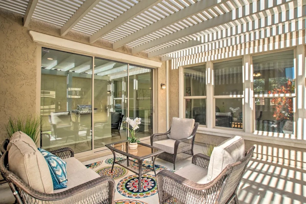 A covered patio area features woven chairs surrounding a small coffee table, with a colorful patterned rug beneath. Large glass doors provide a view into the interior, while sunlight casts striped shadows across the space, creating a bright and inviting atmosphere.