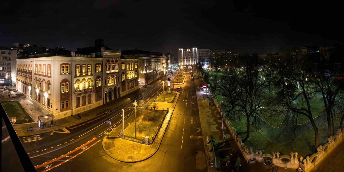 A night view captures a bustling square illuminated by street lights. Historic buildings line the streets, while trees provide a natural element to the scene. The expansive layout highlights the activity of the area, including visible pathways and parked vehicles.