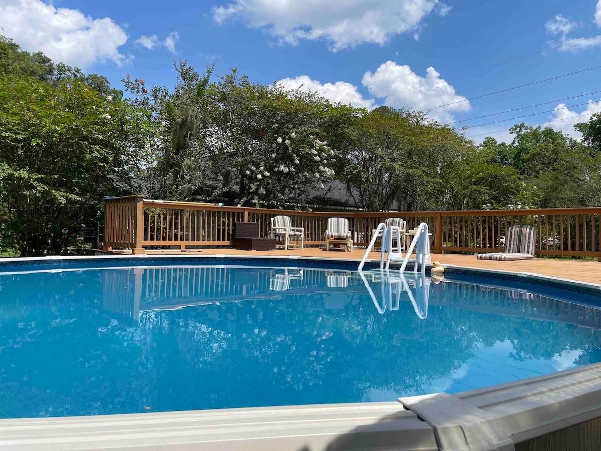 A circular pool is surrounded by a wooden deck, featuring several lounge chairs. Lush greenery and flowering bushes are visible in the background, creating a serene atmosphere under a bright blue sky with fluffy clouds.