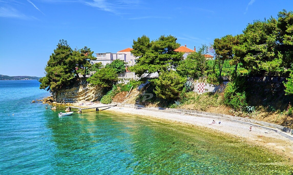 A serene beach scene features a gentle shoreline with clear water, surrounded by lush greenery. A few boats are anchored near the shore. The distant coastal buildings with red rooftops contribute to the calm atmosphere, while the blue sky adds a bright backdrop.