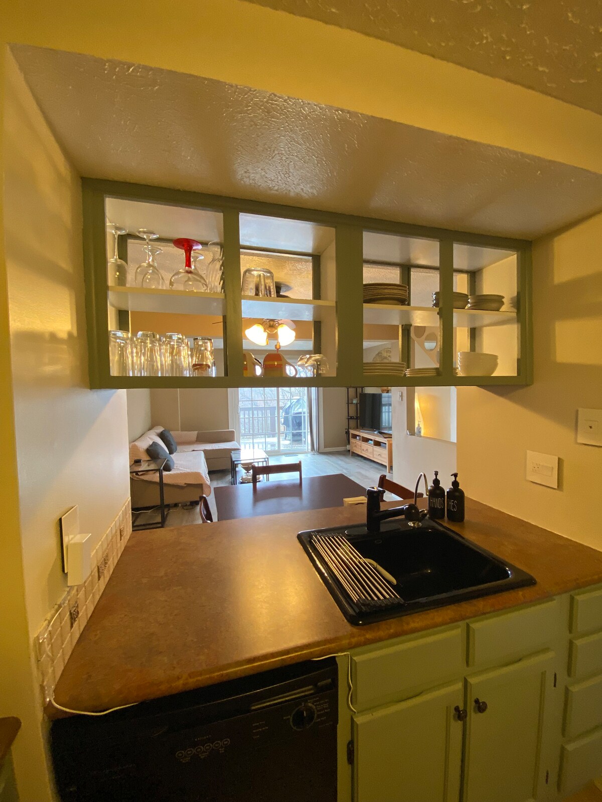 The kitchen area features a well-organized layout with a dark countertop and a black sink. Above, glass-front cabinets display various kitchenware, including glasses and dishes. The space opens into a cozy living area, illuminated by natural light coming from nearby windows.