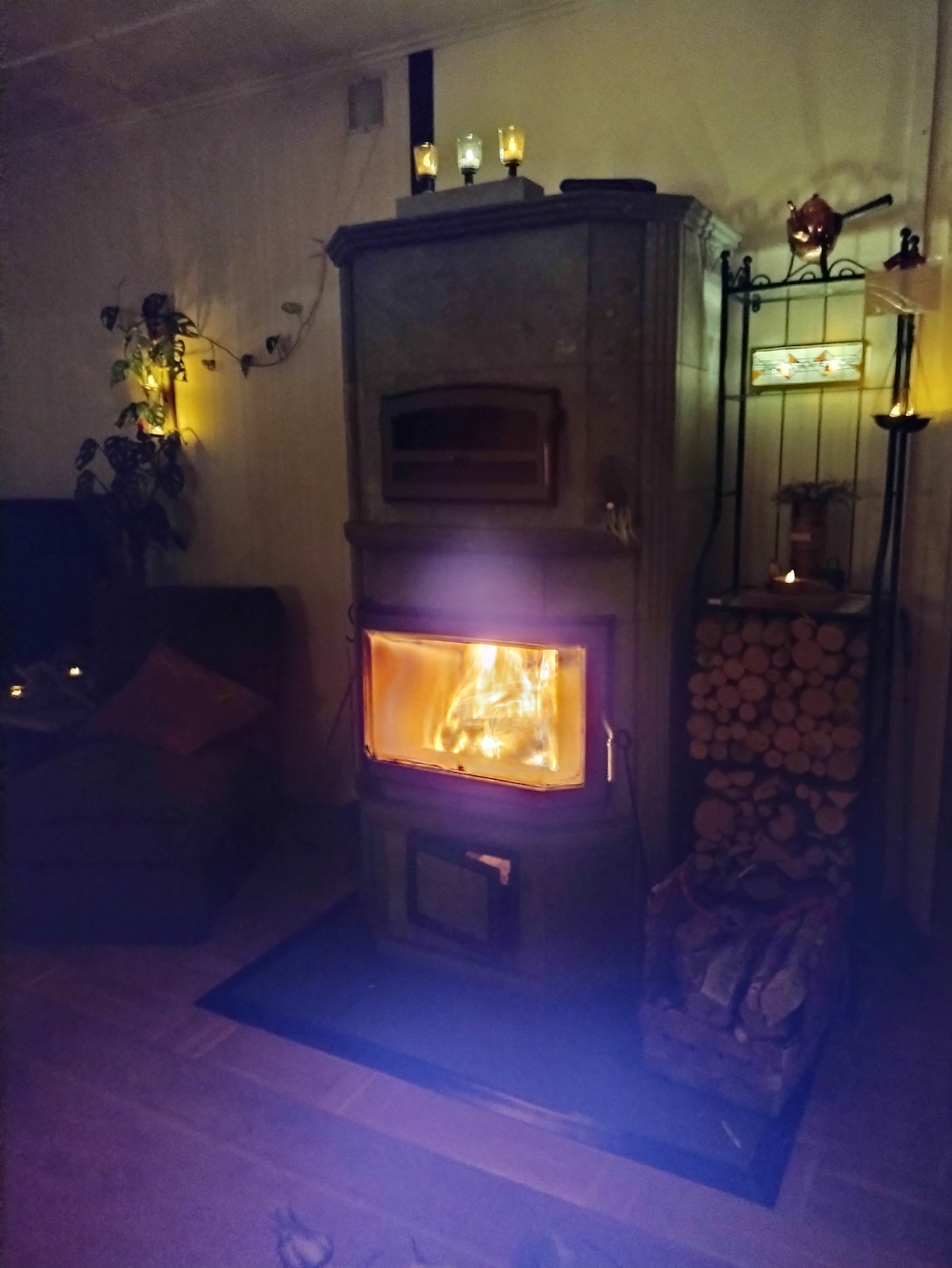 A traditional wood-burning stove is positioned prominently, with flames visible through the glass door. Surrounding the stove are decorative elements, including candles and plants, which contribute to a cozy atmosphere. A stack of firewood is neatly arranged beside the stove.