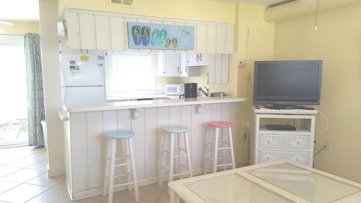 The kitchen area features a light-colored countertop with three pastel-colored stools positioned at the breakfast bar. Essential appliances, including a refrigerator and microwave, are neatly arranged. A flat-screen TV is set on a white unit, and soft natural light enters through a nearby window.
