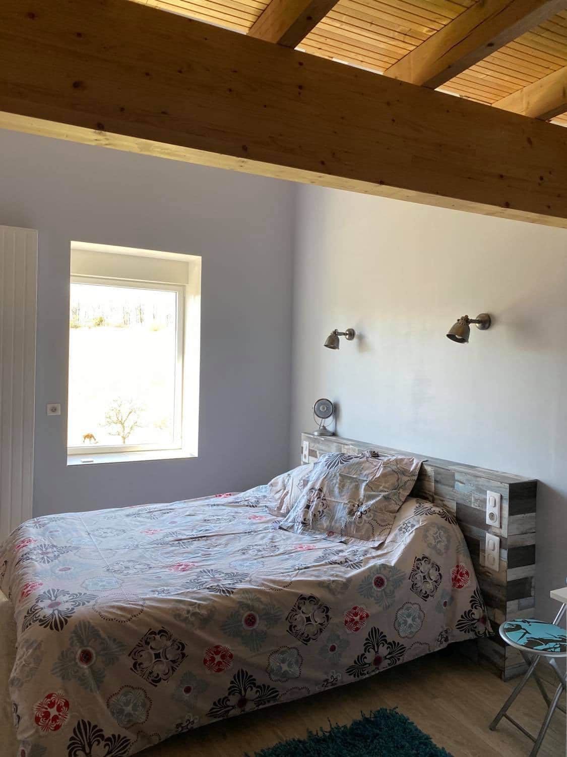 A light-filled bedroom features a spacious bed dressed in a patterned coverlet. Soft lighting from wall sconces complements the natural light streaming through a window. The room's design reflects a blend of rustic and modern elements, with wooden beams enhancing the sense of openness.