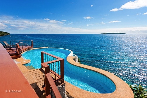 An infinity pool extends over a cliff, surrounded by a smooth stone deck. The vibrant blue water seamlessly merges with the ocean below, reflecting the clear sky. Beyond, a distant island can be seen on the horizon, contributing to the serene coastal view.