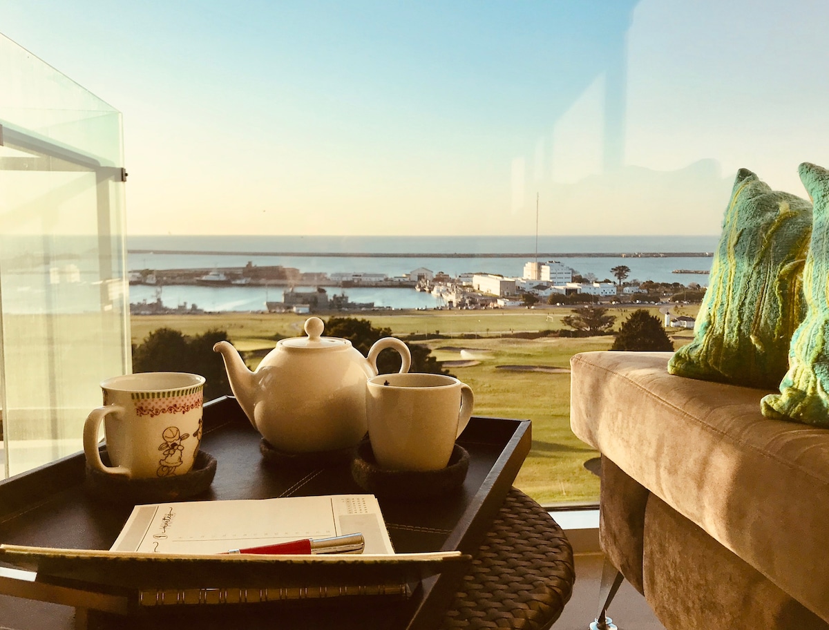 A tranquil scene captures a balcony with a view of the ocean and coastal landscape. On a woven table, a teapot, two cups, and a notepad are arranged. Soft cushions are placed on a nearby couch, inviting relaxation while enjoying the scenery.