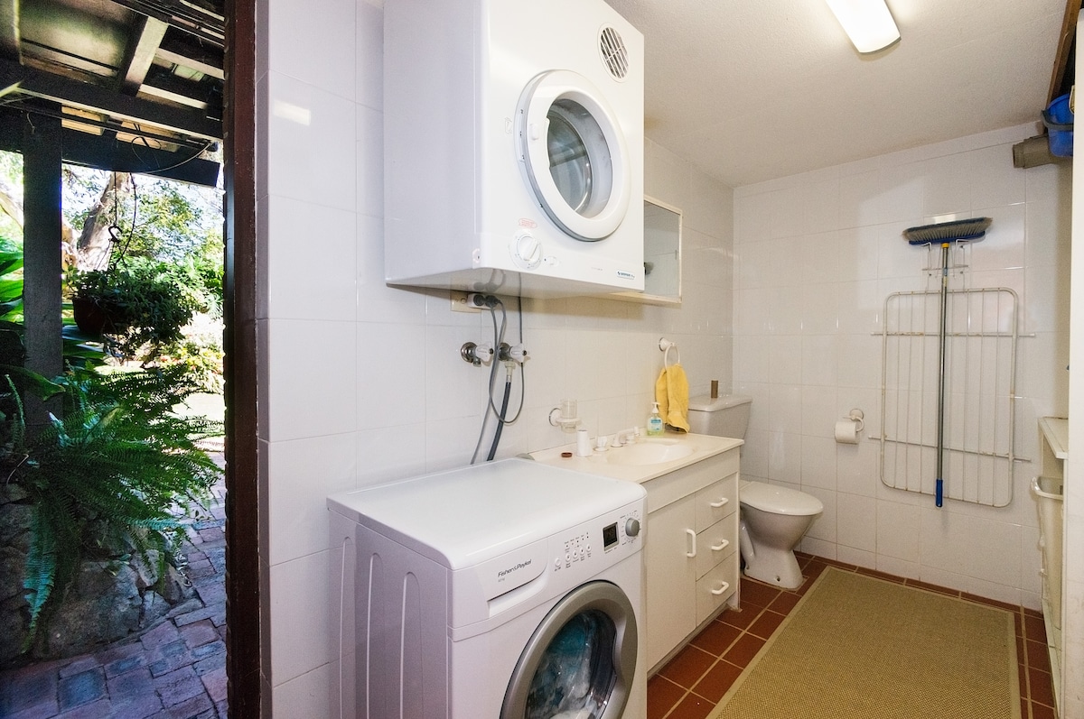 A functional laundry area features a stacked washer and dryer against a clean white wall. A small sink and a toilet are present, while a drying rack is mounted on the wall. Natural light filters in from an open door, showcasing greenery outside.
