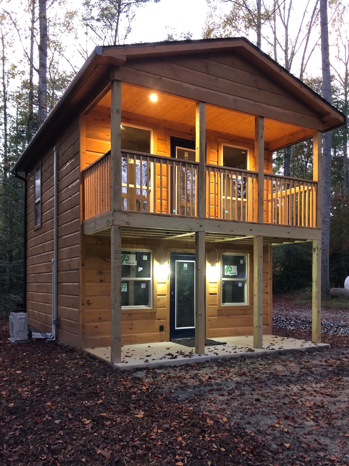 The exterior of the cabin showcases a charming wooden facade, featuring a spacious balcony with wooden railings. Warm lights illuminate the entryway, creating a welcoming ambiance in the forested surroundings. A grassy area is visible in front of the cabin, emphasizing the natural setting.