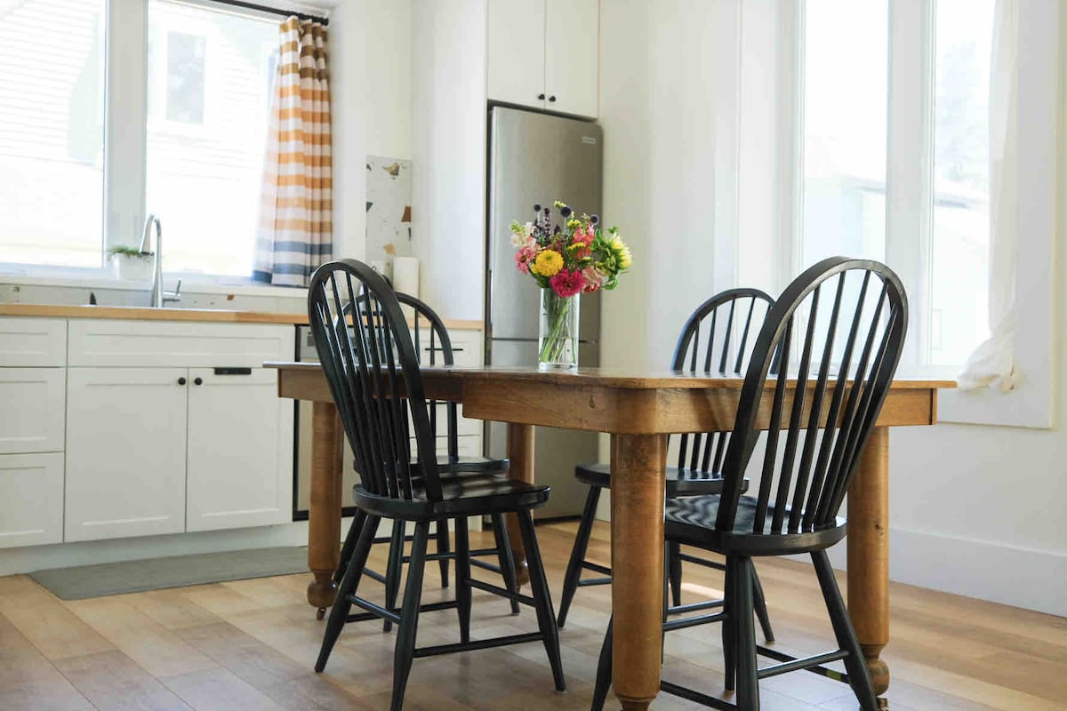 A bright dining area features a wooden table surrounded by four black chairs. A vase filled with colorful flowers sits at the center of the table. Natural light floods the space through large windows, complemented by striped curtains in warm tones.