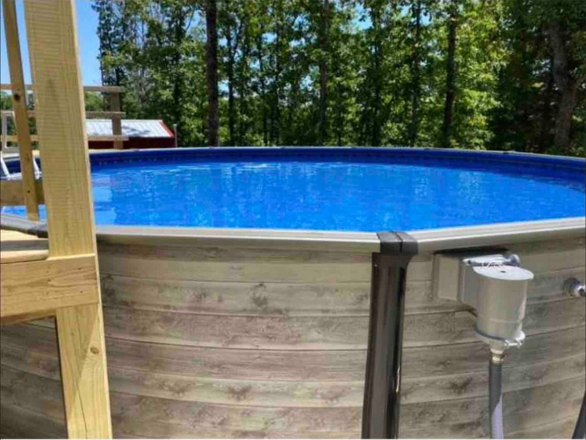 A round, above-ground swimming pool is surrounded by wooden decking. The clear blue water reflects sunlight, while trees in the background provide a natural setting. The pool features a smooth, light-colored exterior made of wood-like material.
