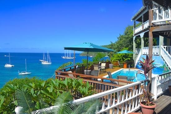 A spacious deck surrounds a pool, offering views of the turquoise water and anchored boats in the distance. Green umbrellas provide shade over outdoor seating arrangements. Lush tropical plants border the area, enhancing the serene, coastal setting of this Caribbean villa.