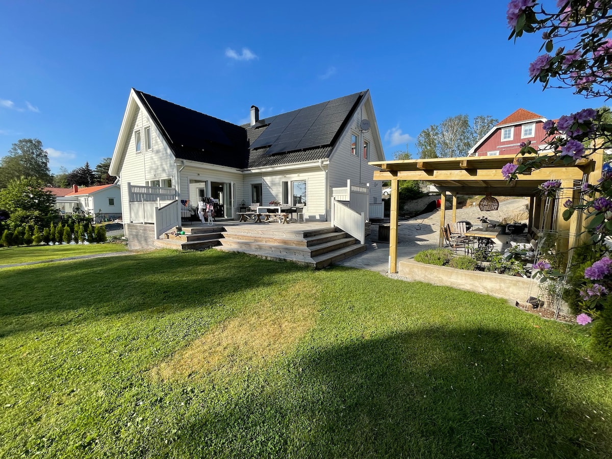 A two-story house is shown, featuring a sloped roof and a large deck with outdoor seating. A pergola with a dining setup is visible in the garden, surrounded by well-maintained grass and blooming flowers. Clear skies indicate pleasant weather.