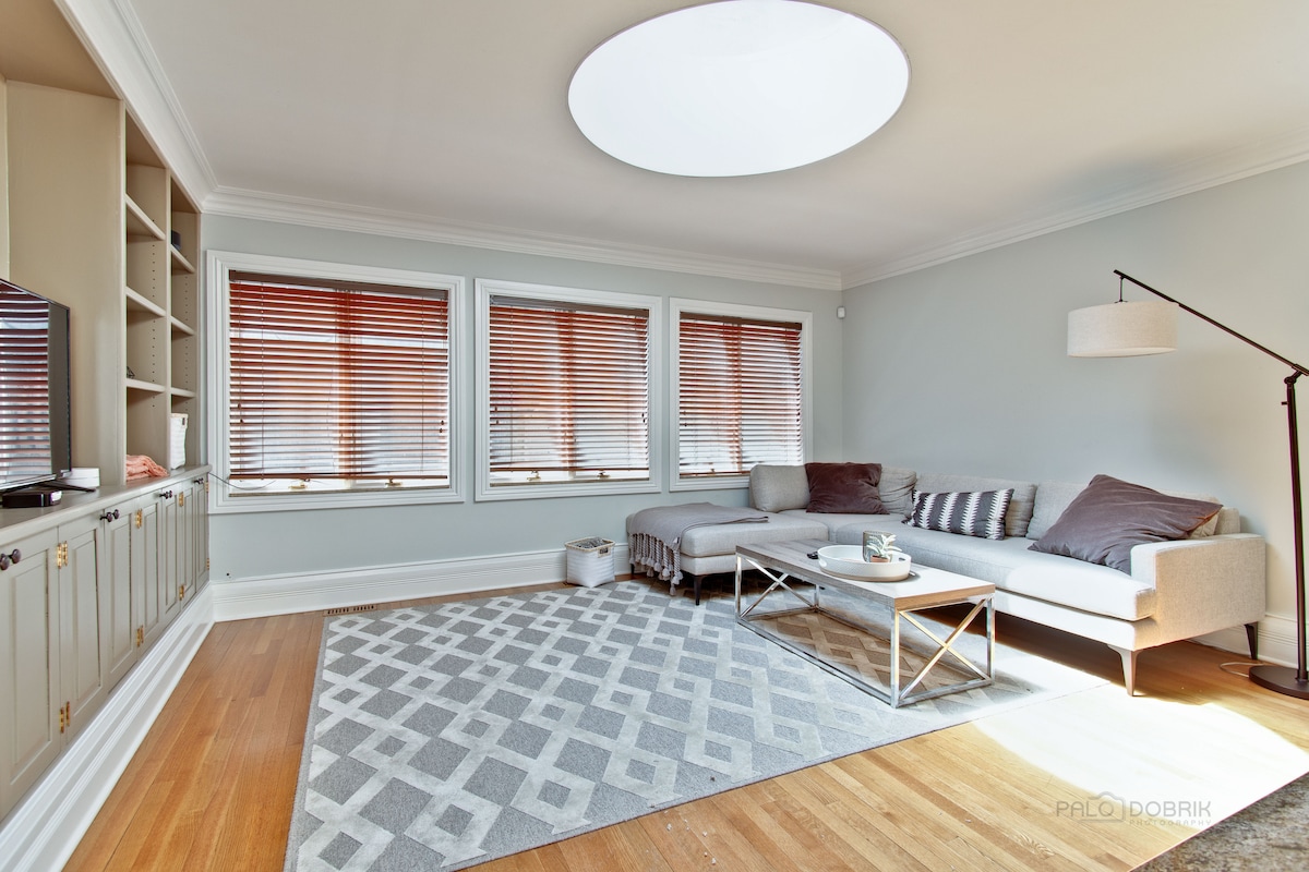 A spacious living room is highlighted by a large circular skylight, allowing natural light to fill the area. Comfortable seating is arranged on a patterned rug, with a modern coffee table at the center. Window shades partially cover the three windows, adding a touch of privacy.