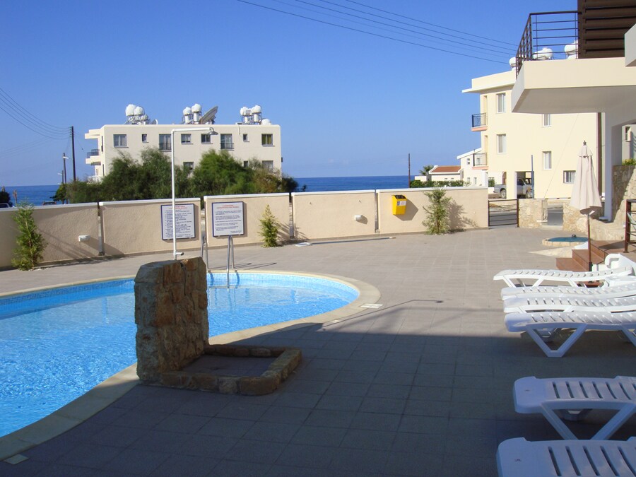 A tranquil swimming pool is surrounded by tiled decking, with sun loungers positioned nearby. Towering buildings and greenery frame the area, while the distant sea is visible under a clear blue sky. Informational signs are placed near the pool, adding to the inviting atmosphere.