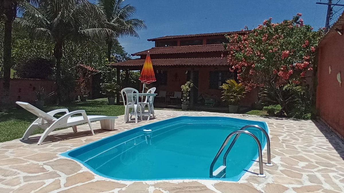 A clear blue swimming pool is framed by a stone patio, with a white lounge chair positioned nearby. A table and chairs sit under a vibrant umbrella, with lush greenery and flowering plants surrounding the area. The house is visible in the background, highlighted by red roof tiles.