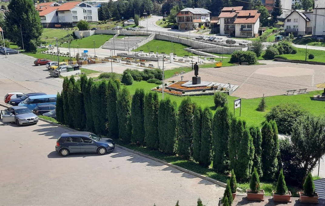 A view of a neatly landscaped area featuring manicured hedges and a statue at the center. Surrounding trees provide shade, while nearby buildings and a parking area are visible in the background, enhancing the open atmosphere of the space.