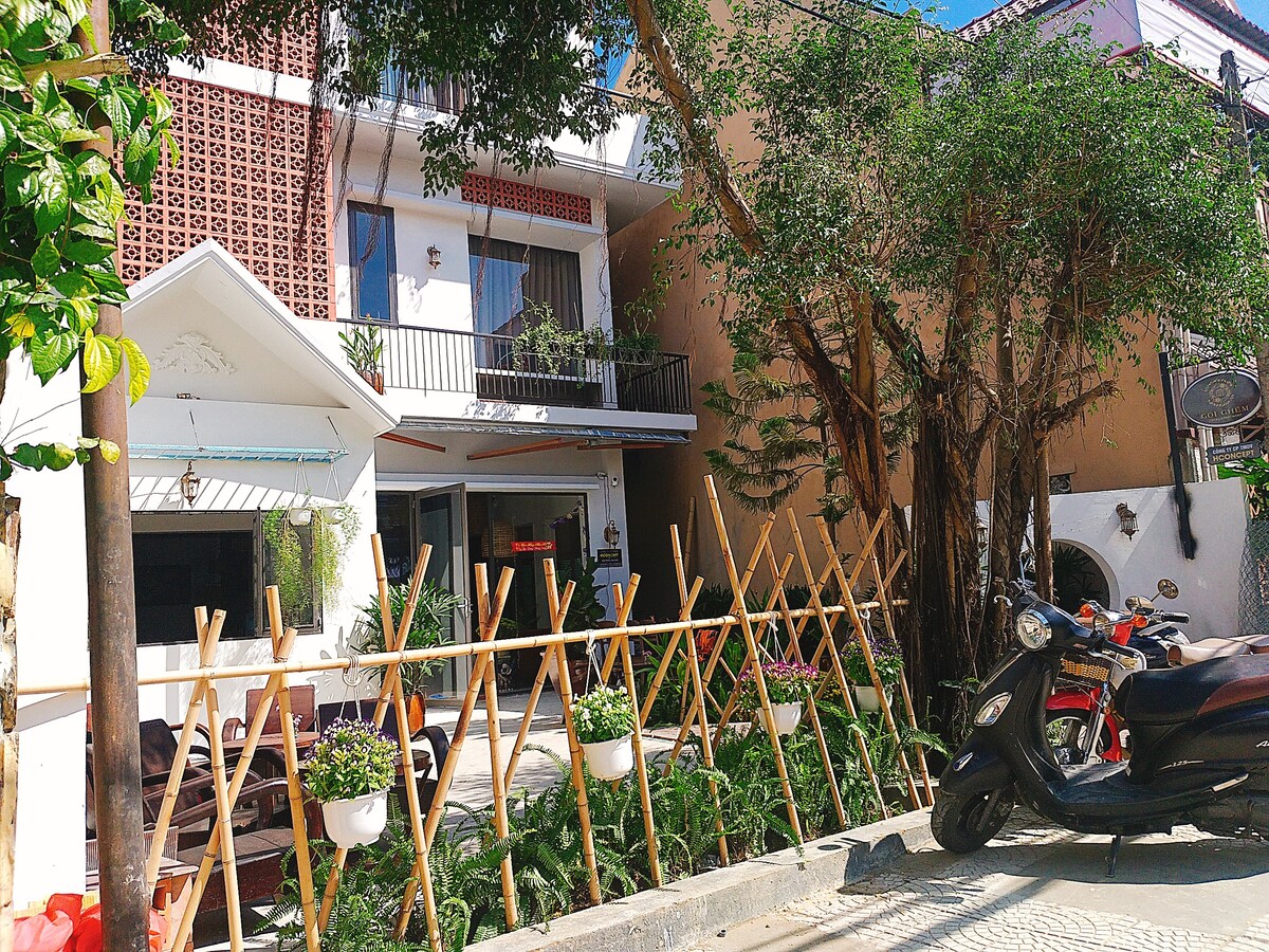 The exterior features a welcoming facade with a blend of modern design and rustic charm. A bamboo fence surrounds a green garden area, while potted plants add life. A parked motorcycle is visible, indicating accessibility nearby.