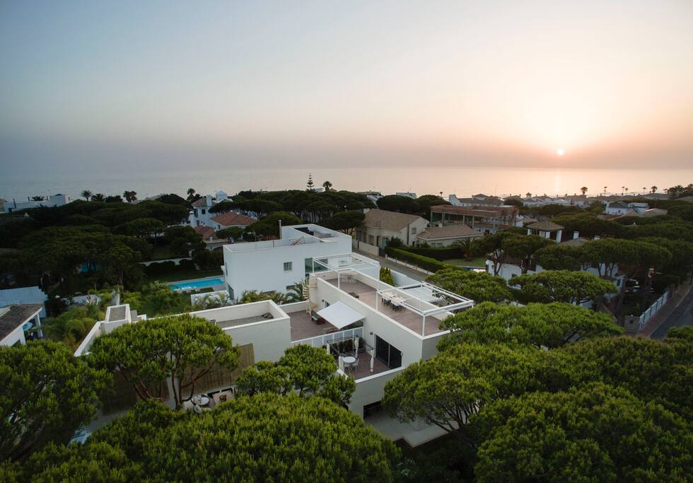 Vista aérea de nuestra casa en la que podéis apreciar muy bien sus terrazas de madera y espacios exteriores protegidos por toldos eléctricos, pérgolas y toldos de gran tamaño y el increíble entorno en medio de un pinar protegido con palmeras y exótica vegetación y el mar al fondo. El mar está tan cerca que puedes bajar descalzo a la playa y desde la casa se escucha y se huele sin los inconvenientes y el jaleo de estar en primera linea.