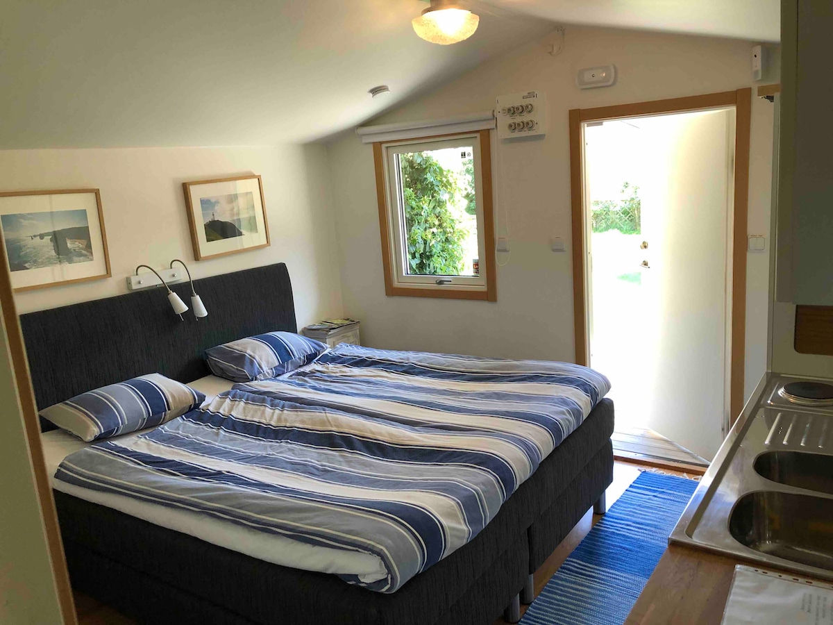 A comfortable sleeping area is presented with a spacious bed layered in striped bedding. Natural light enters through a nearby window and an open door leads to the garden. Framed pictures adorn the walls, complementing the minimalist decor.
