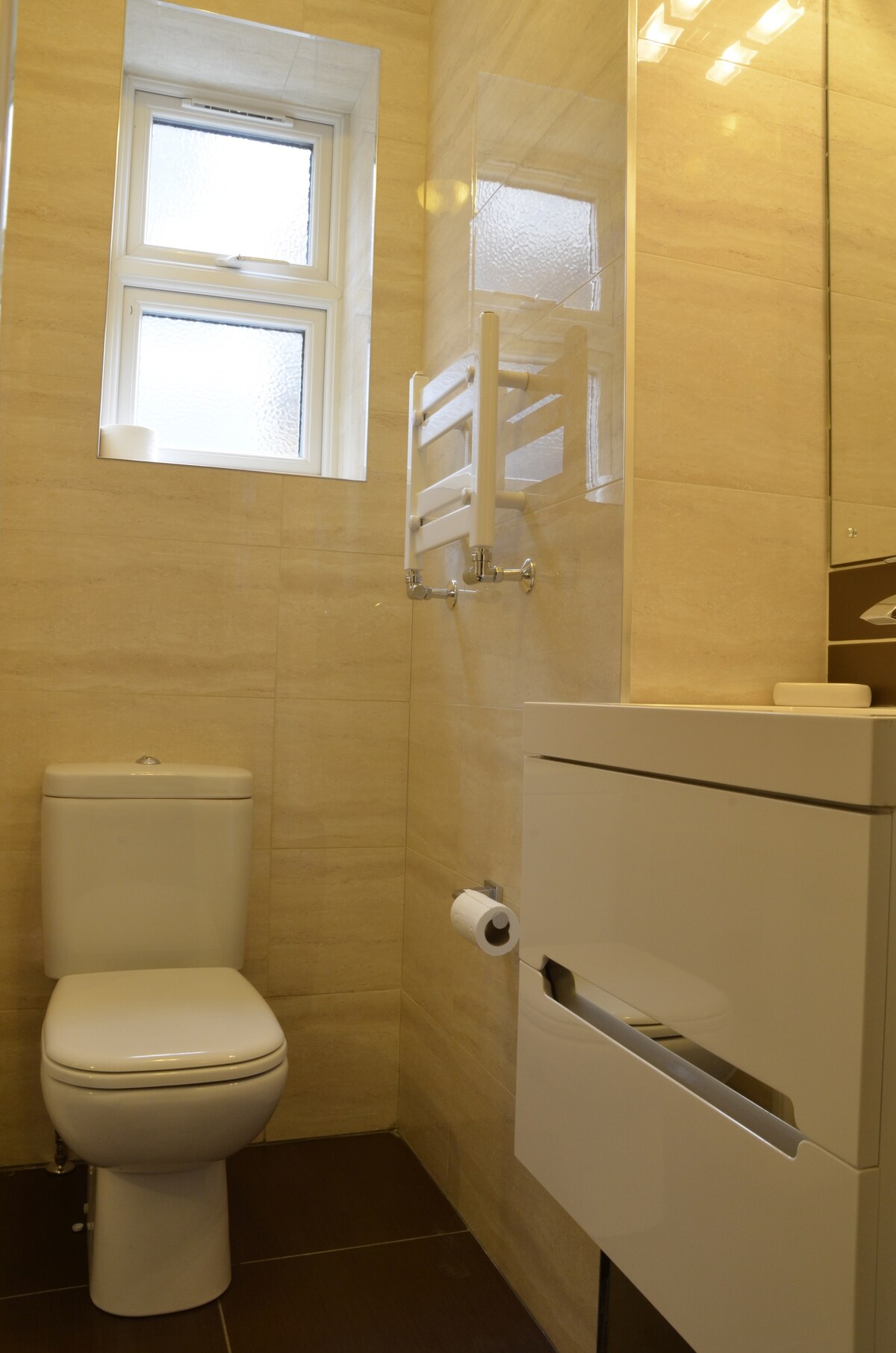A compact en-suite bathroom is presented, featuring a modern toilet and a sleek vanity unit with a storage drawer. Natural light filters through a small window, illuminating the neutral-toned tiles that cover the walls and floor. A towel heater is mounted on the wall for added convenience.