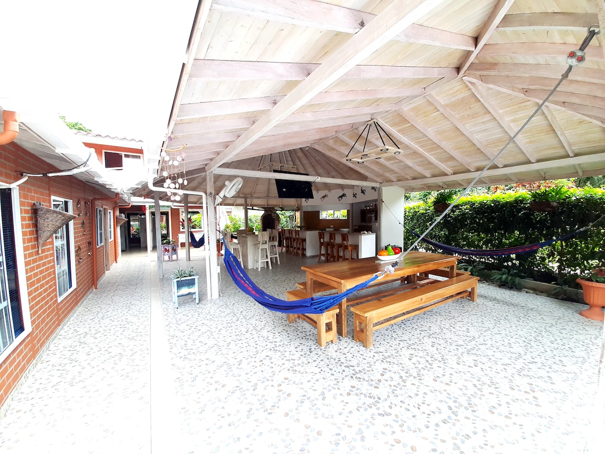 A spacious common area features a large wooden dining table surrounded by comfortable seating and decorative plants. Hammocks are installed for relaxation, with a ceiling decorated in light wood. Natural light enhances the open space, which is designed for social gatherings.