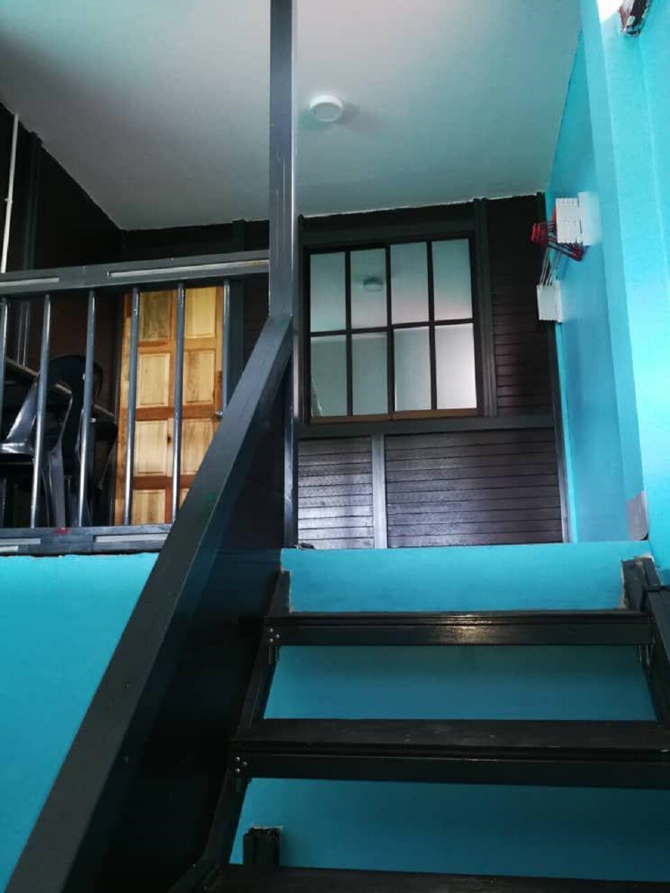A staircase with black metal steps leads upwards, flanked by teal walls. A door with a wooden frame and square window panels is visible at the top of the stairs. The area features a minimalist design with a combination of light and dark wood accents.