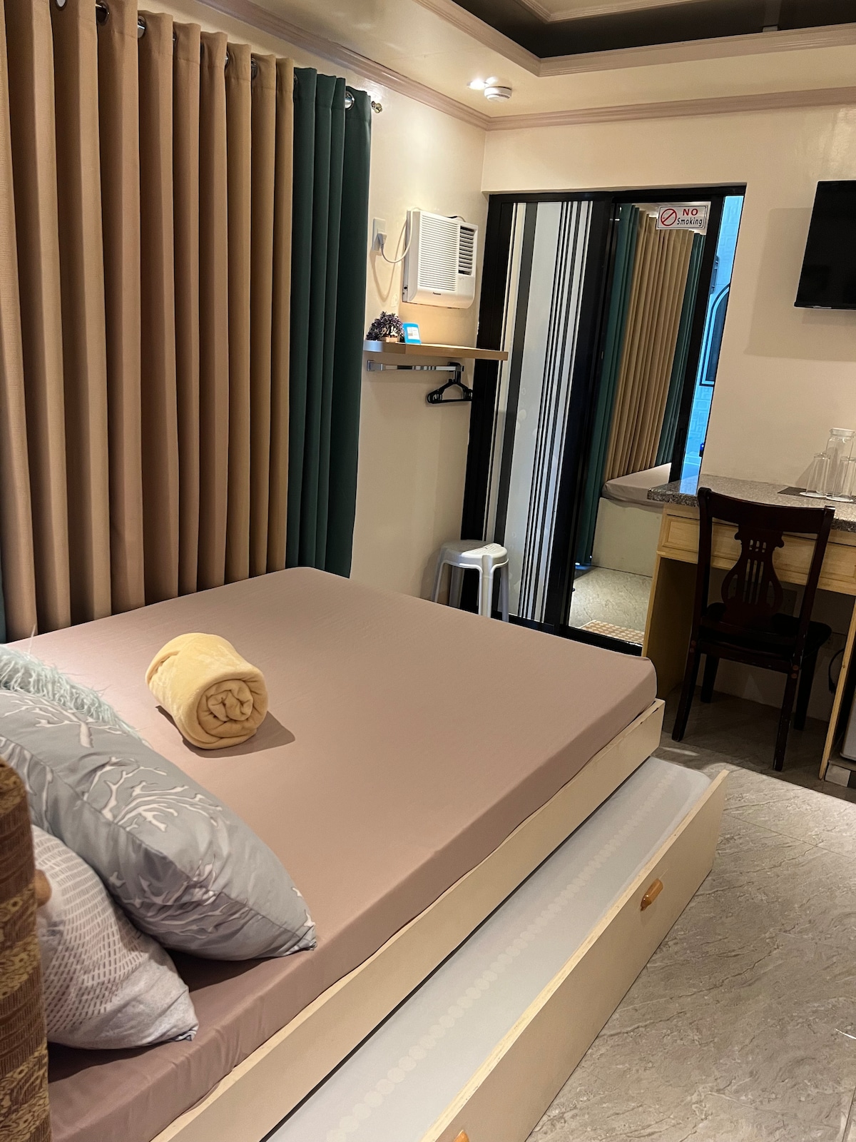 A well-appointed bedroom features a spacious bed with a neutral-colored mattress and decorative pillows. Light-colored curtains frame a large mirror, and a small desk with a chair is positioned nearby. An air conditioning unit is mounted on the wall, adding comfort to the space.