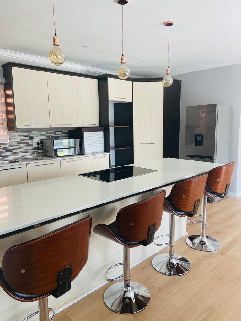 A modern kitchen features a sleek island with four high-backed bar stools. Cabinets in muted tones are complemented by stainless steel appliances and a subtle patterned backsplash. Warm light fixtures hang above, highlighting the functional layout and clear space for cooking and entertaining.
