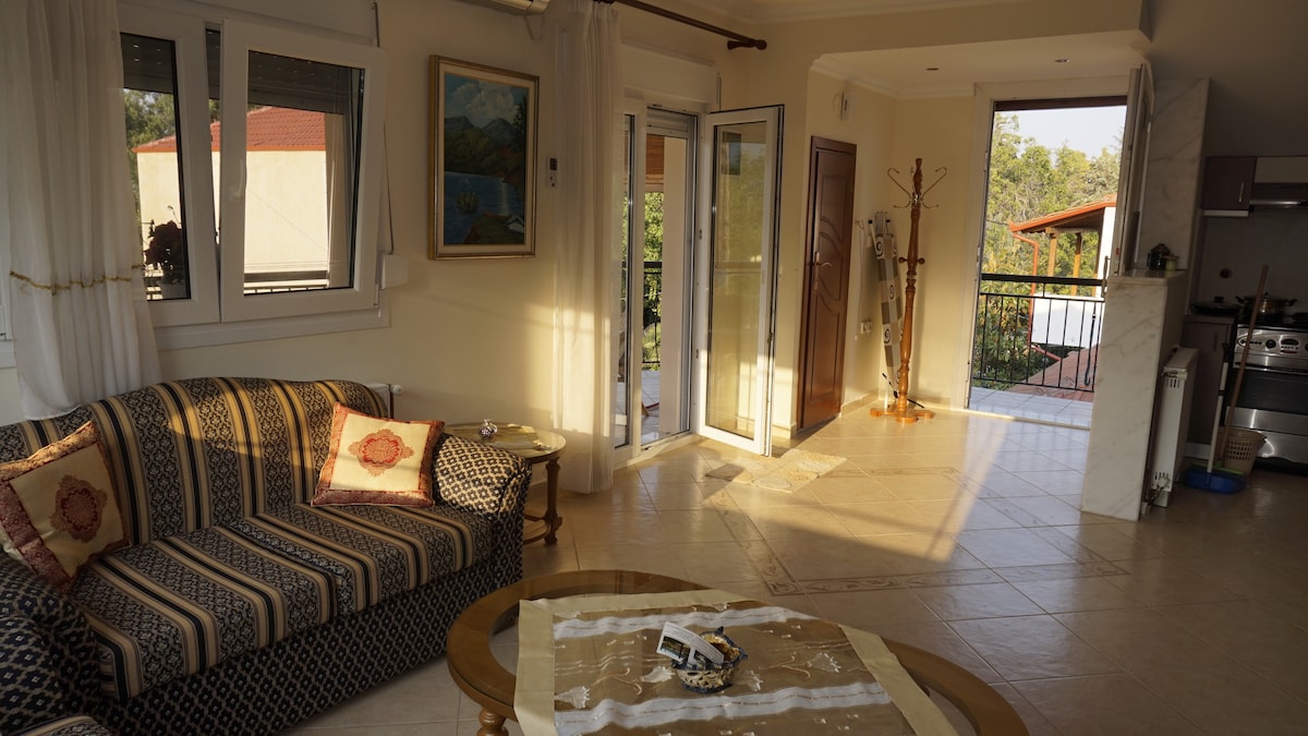 A spacious living area features a patterned sofa with decorative cushions, complemented by a wooden coffee table. Large glass doors open to a balcony, allowing natural light to fill the room. Soft curtains frame the windows, enhancing the serene atmosphere.