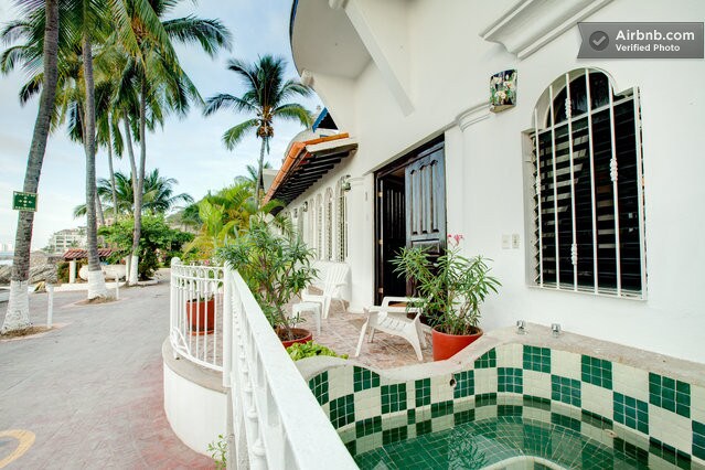 A welcoming entrance is framed by palm trees, with a private jacuzzi visible on the left. Potted plants enhance the outdoor space, while a few seating chairs are placed on the patio. The main door opens into the apartment, bathed in natural light.