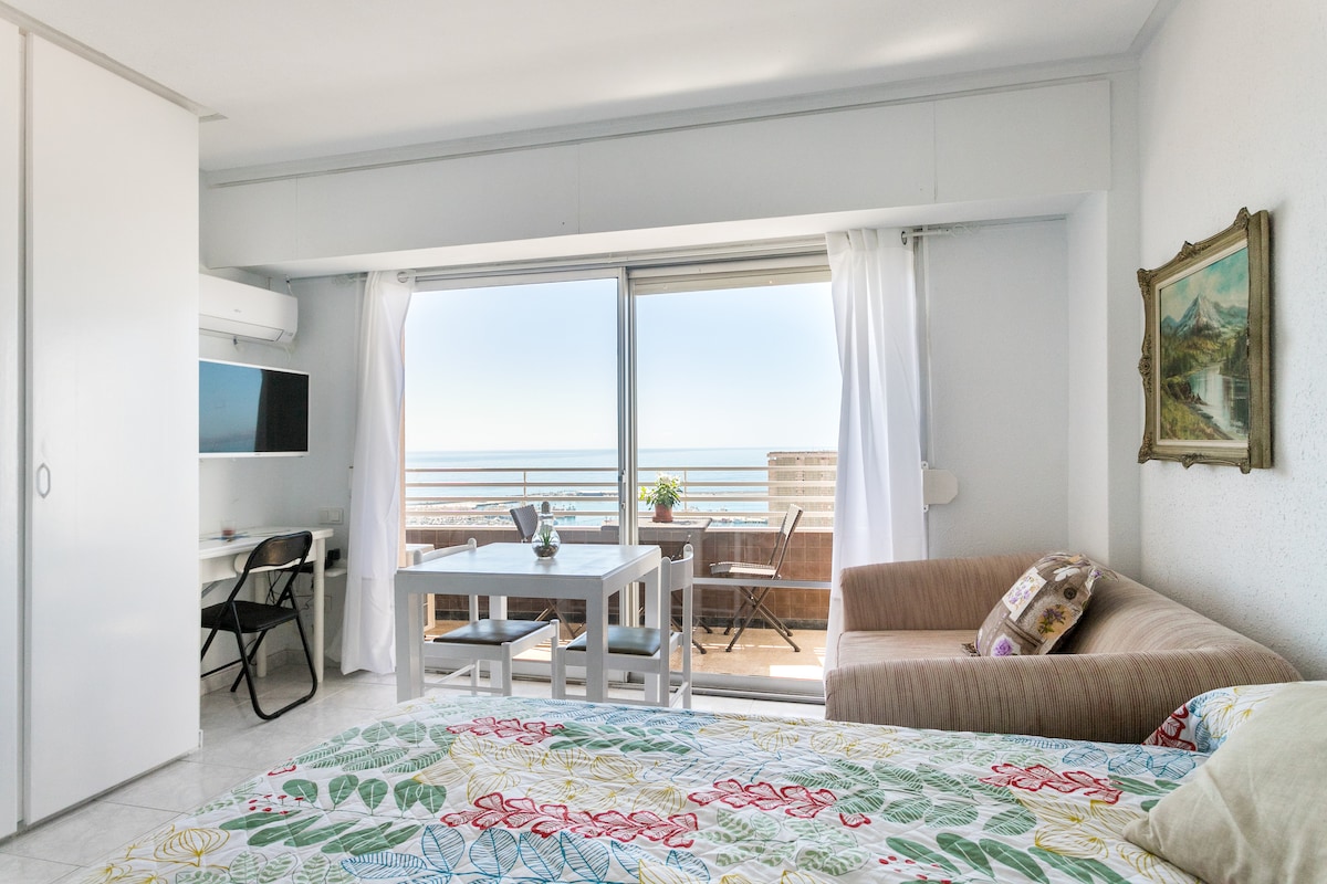 A bright studio space features a spacious bed adorned with a colorful floral quilt. Large sliding glass doors provide stunning views of the Mediterranean and access to a balcony. A dining table and chairs are positioned next to the window, enhancing the usable living area.