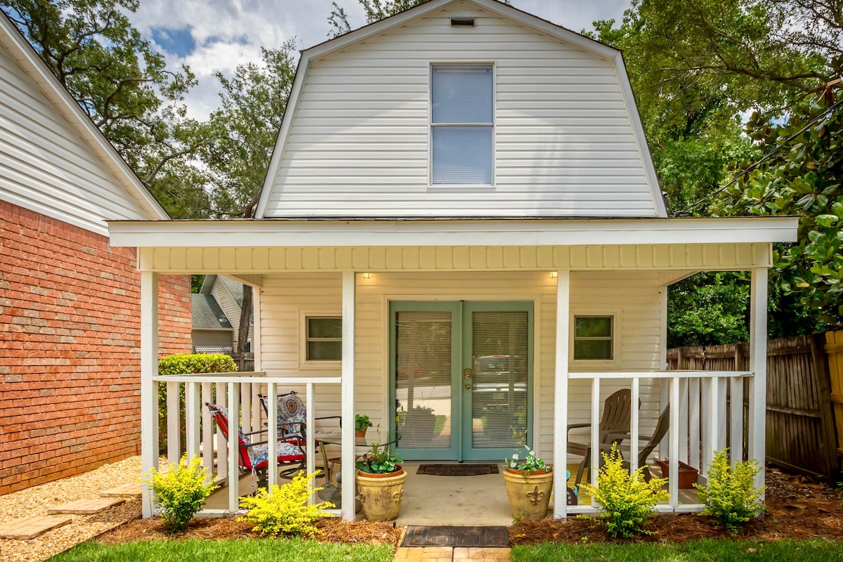 A quaint exterior of a beach-style home is showcased, featuring a welcoming porch with two chairs and potted plants. The building is painted white with a sloped roof and a large central window. Lush greenery surrounds the entryway, adding a refreshing touch.