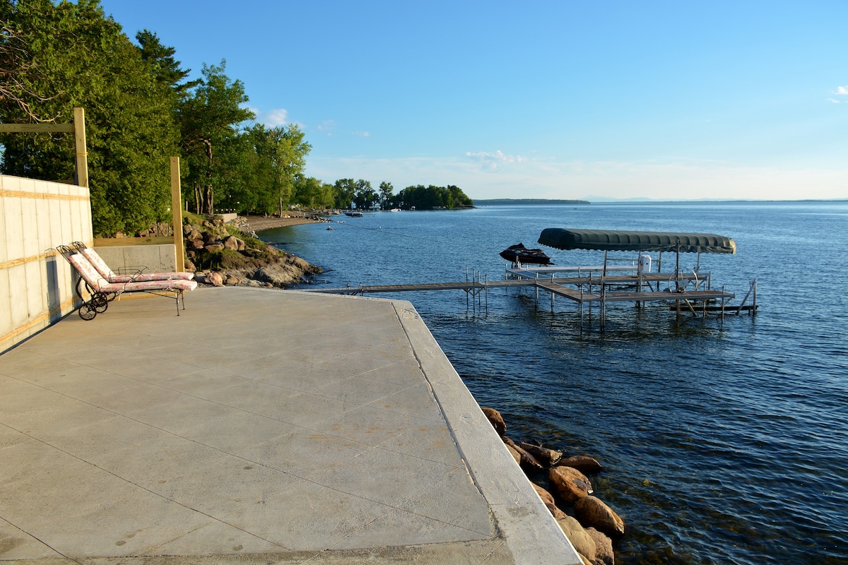 A waterfront patio is visible, featuring a spacious concrete area with two lounge chairs positioned for relaxation. A dock extends into the lake, accommodating a covered boat lift. The calm water reflects the clear blue sky and is surrounded by greenery along the shore.