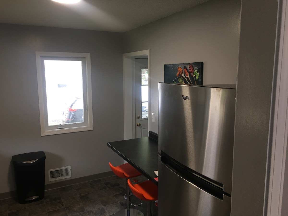 A modern kitchen area features a stainless steel refrigerator and a sleek, dark countertop with orange bar stools. A window provides natural light and a view of the outside. A door leads to another room, enhancing the space's accessibility.