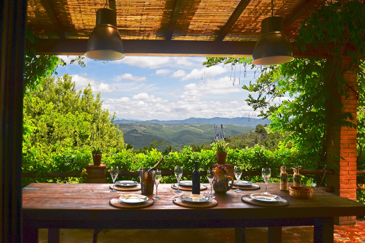 An outdoor dining area is set with a wooden table, awaiting guests. Elegant tableware complements a scenic view of rolling hills and greenery. Soft lighting from hanging fixtures enhances the natural ambiance, while lush plants frame the space, creating an inviting atmosphere for dining amidst nature.