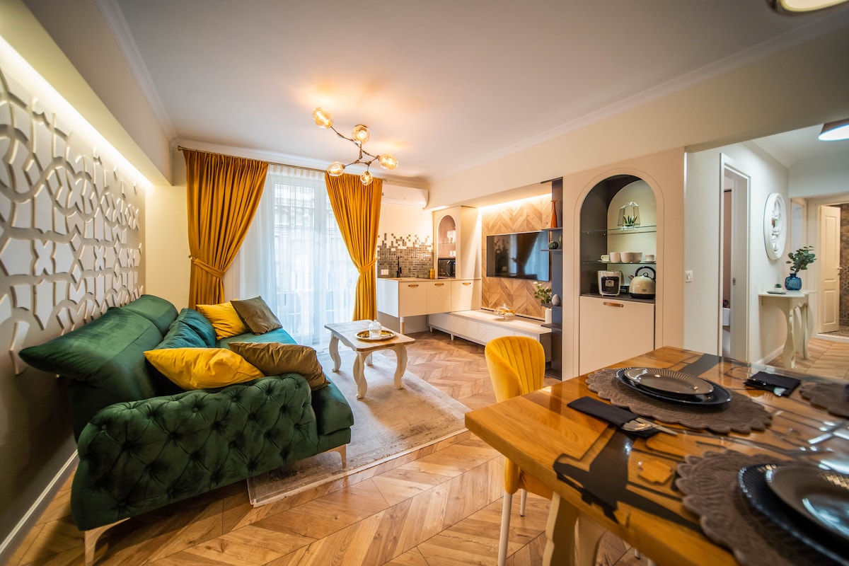 The living area features a cozy green sofa adorned with yellow cushions, paired with a wooden coffee table. A stylish entertainment unit displays a television, and large windows are dressed in golden curtains, allowing natural light to fill the space. A dining table is set nearby.
