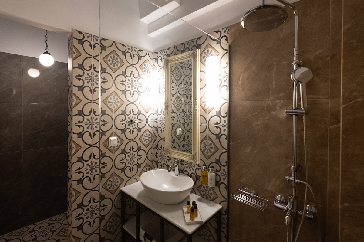 A modern bathroom features decorative tiles with intricate patterns on the walls. A circular sink rests on a marble countertop, complemented by a framed mirror. Shower fixtures are elegantly arranged, with overhead lighting providing a warm glow throughout the space.
