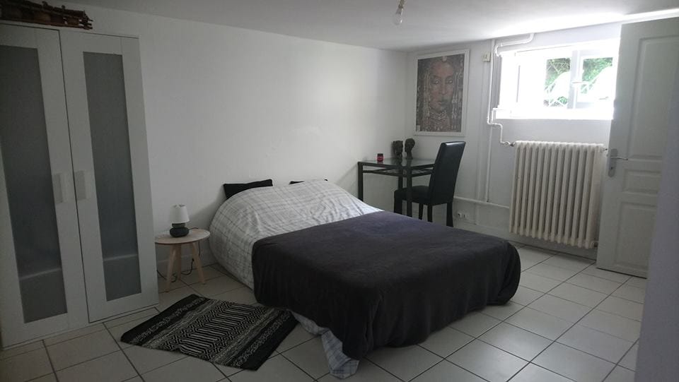A well-appointed bedroom is visible, featuring a spacious bed covered with a dark blanket. A bedside lamp rests on a small table beside the bed. A chair and table are positioned against the wall, and a tall cabinet with glass doors is present. Light enters from a nearby window.