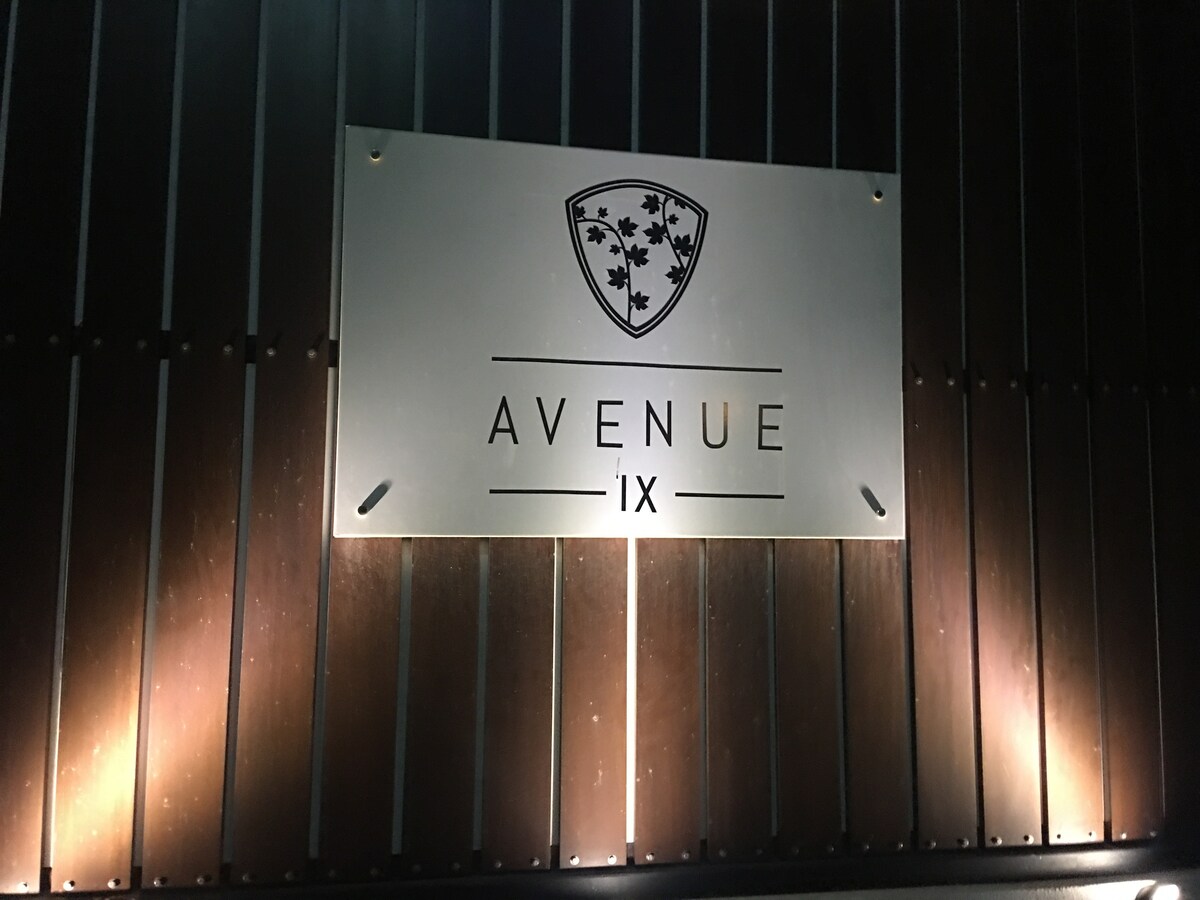 A sleek metal sign displaying the name 'Avenue IX' is illuminated against a dark wooden background. The sign features a logo with shield design and decorative leaves, enhancing the modern entrance's aesthetic.