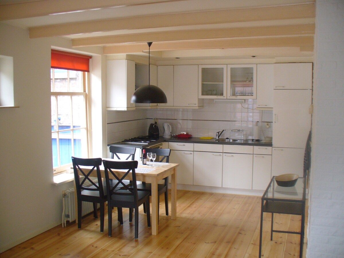 A compact kitchen area is equipped with modern appliances, including a stove and refrigerator. A small dining table with black chairs sits nearby. Natural light filters through a window with a bright orange shade, highlighting the wooden floor and sleek cabinetry.