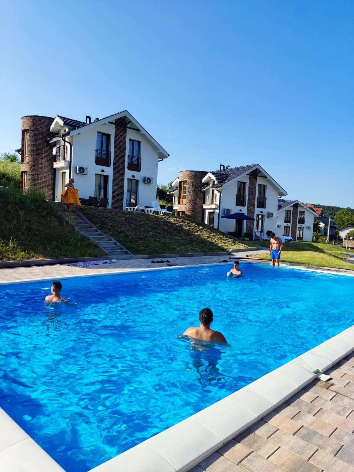 A shared swimming pool is surrounded by well-maintained green space and a row of stylish two-story buildings. Several guests are enjoying the pool, with bright blue water reflecting the clear sky. Lounge chairs are positioned nearby, offering comfortable seating.