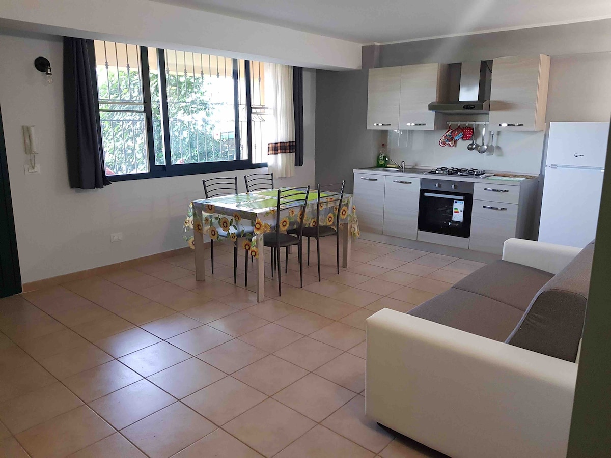 A well-lit kitchen and dining area features a modern dining table with four chairs, a refrigerator, and a gas stove. A comfortable sofa is positioned nearby. Natural light flows through large windows with vertical blinds, offering views of the garden outside.