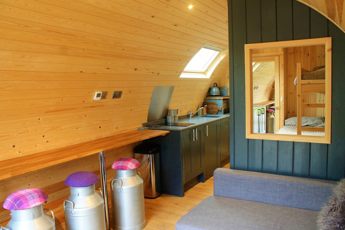 A compact kitchen space includes wooden countertops and modern appliances. Two silver milk jugs are featured, with vibrant cushions on three bar stools. A cozy seating area is visible, and a mirror reflects the light from a nearby skylight, enhancing the openness of the cabin.