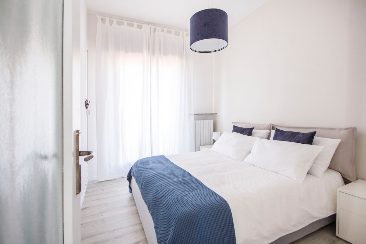 A bright bedroom features a spacious bed with white linens and decorative pillows in shades of blue. Soft light filters through sheer white curtains. A simple pendant lamp hangs above the bed, and light-colored flooring adds to the airy feel of the room.