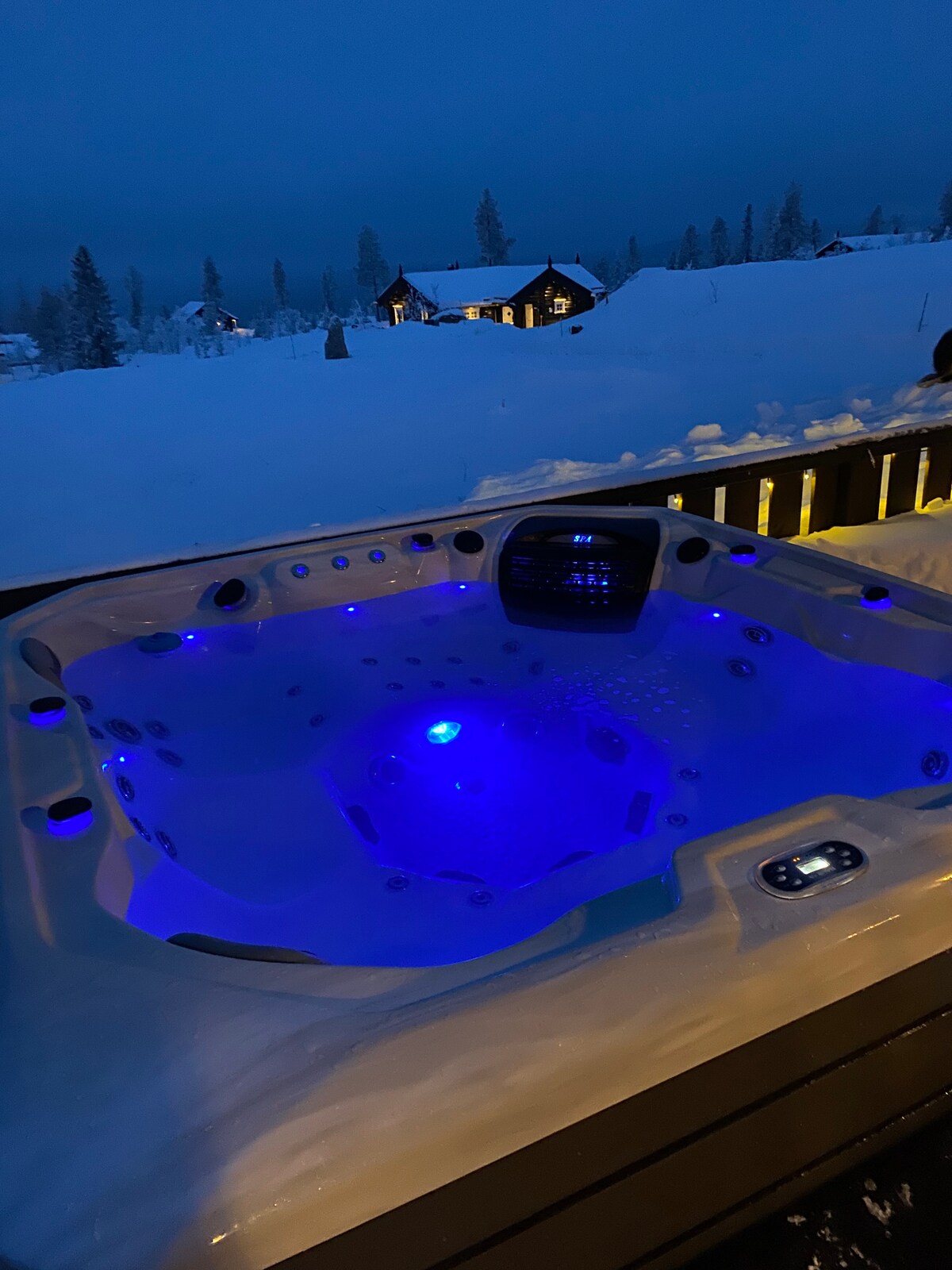 A new Jacuzzi is surrounded by winter scenery, with soft blue lights illuminating the water. Snow blankets the adjacent area, enhancing the tranquil atmosphere. The cabin's silhouette can be seen in the background, creating a serene setting for relaxation.