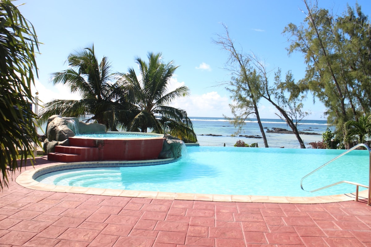 A serene swimming pool overlooks the lagoon, with palm trees framing the view. The pool features a gentle curve and a connected hot tub. The bright blue water reflects the clear sky, and the natural surroundings enhance the tranquil setting.