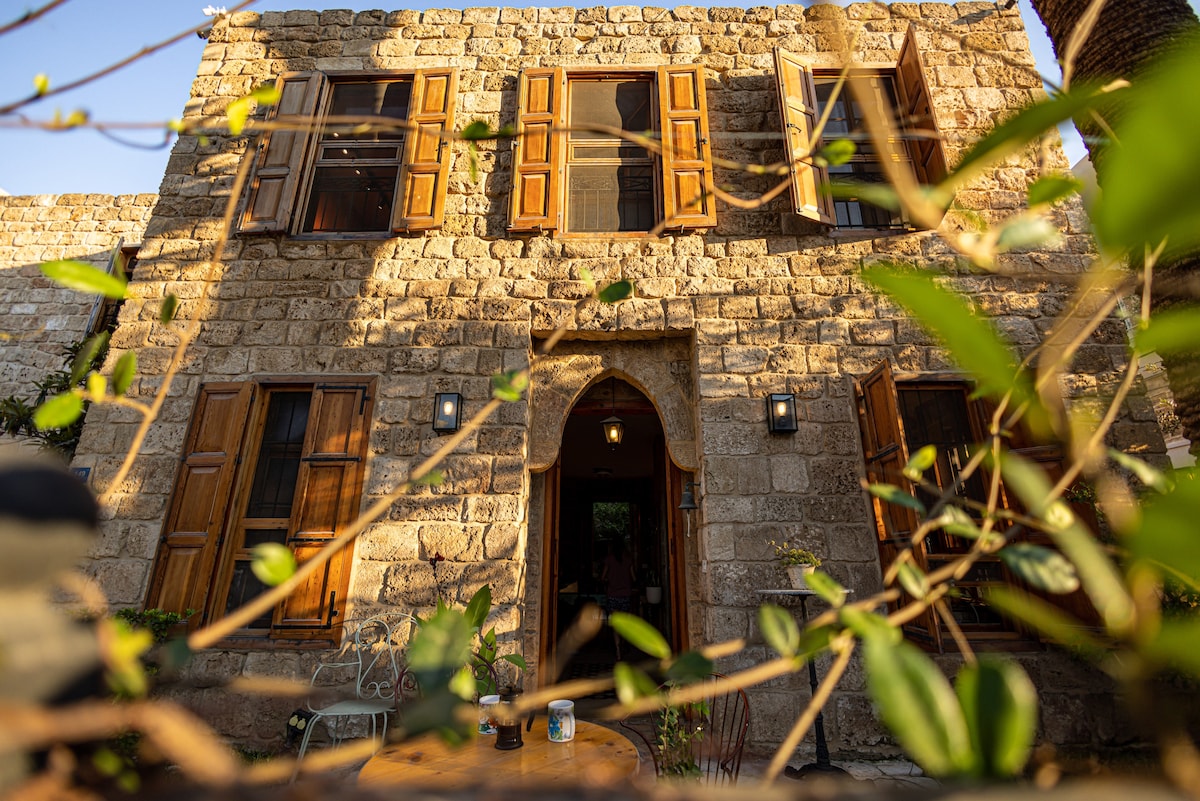 A traditional Lebanese house is showcased, built from stone with warm wooden shutters framing the windows. An arched doorway is centrally positioned, inviting entry. Lush greenery partially obscures the view, enhancing the structure's connection to the surrounding landscape.